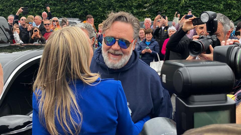 Russell Crowe being greeted by Laura Bonner as he emerges from a car after arriving at the Muff Liquor Company distillery in County Donegal. He is facing towards the camera, smiling at Ms Bonner while wearing sunglasses. She has her back to the camera and is wearing a bright blue jacket. The are surrounded by photographers taking pictures and dozens of onlookers, many of whom are taking photos on camera phones.