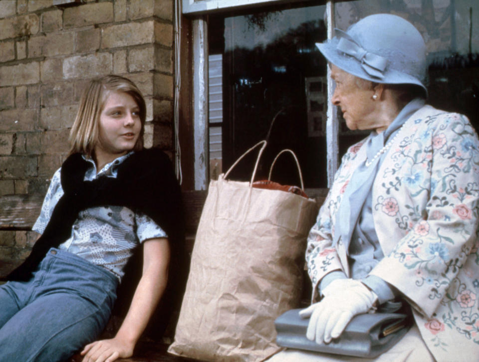 A scene from the movie "Candleshoe" featuring Jodie Foster sitting on a bench next to an elderly woman wearing a floral coat and a hat