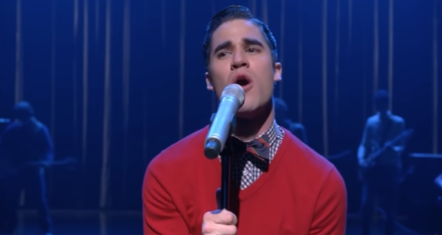 Man performing on stage with microphone, wearing a red sweater vest and tie, with band in the background