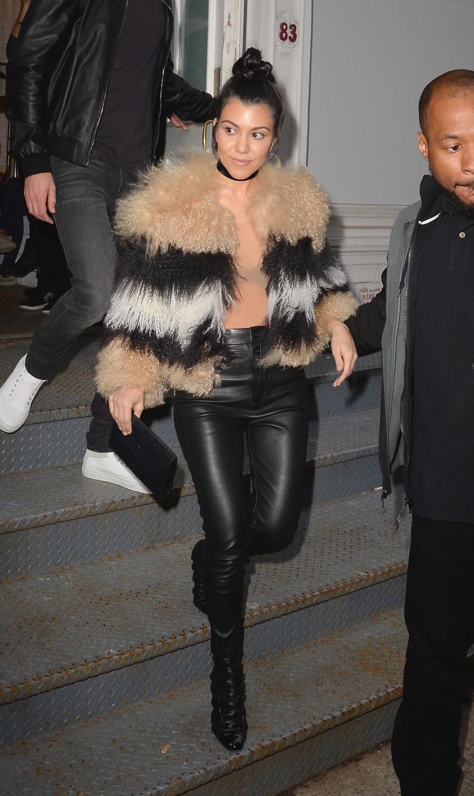 Kourtney in a beige, black, and white feather and fur cropped coat, a sheer top, and leather pants and boots.