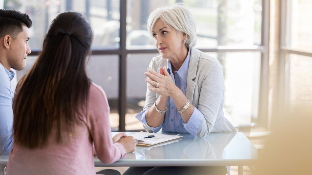 A female mental health professional with gray hair looks intently at a husband and wife who have come to her office to discuss relationship problems.