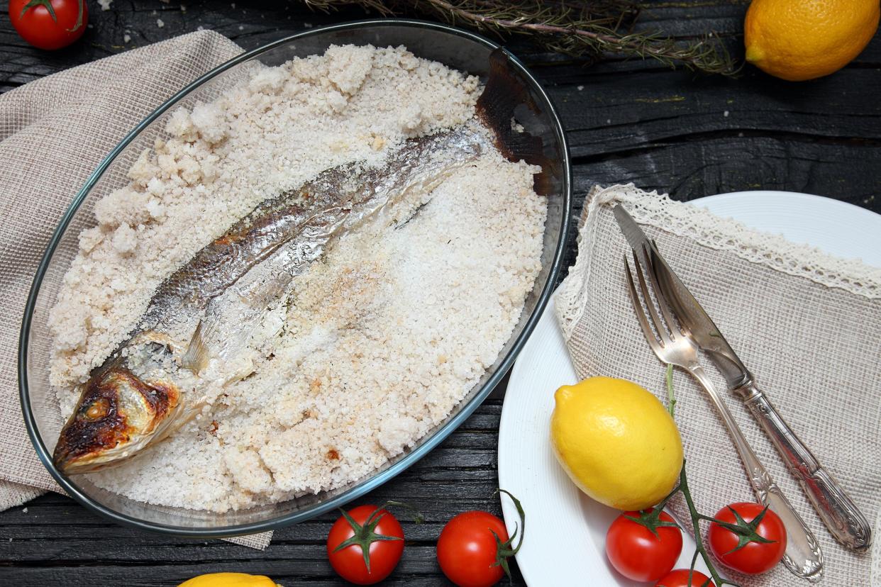 Salt-baked fish in a glass baking bowl with a plate with a fork and knife surrounded by ingredients