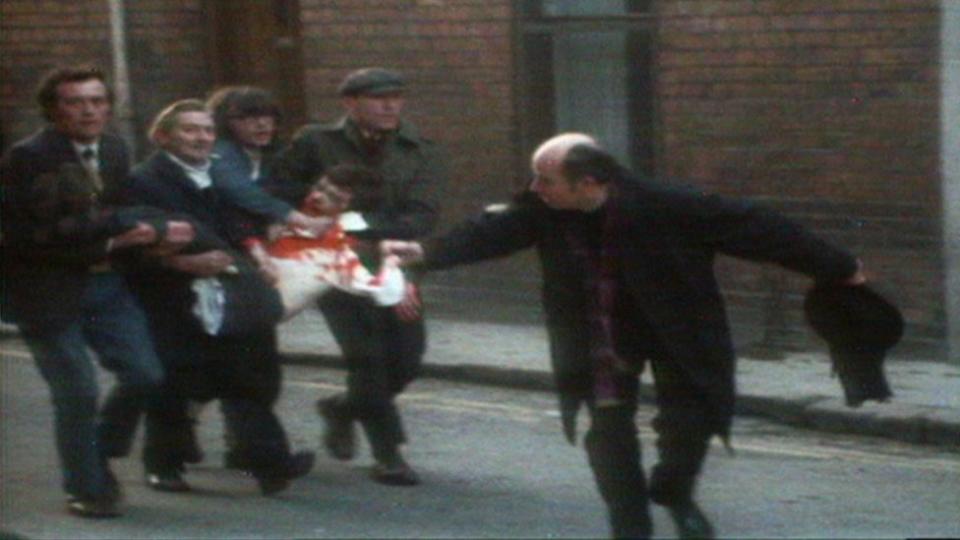 Un sacerdote agita un pañuelo manchado de sangre mientras cuatro hombres llevan a un hombre herido y ensangrentado por las calles de Londonderry durante The Troubles.