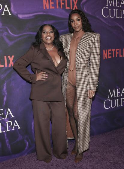 Sherri Shepherd in a brown suit standing next to Kelly Rowland in a long brown, checkered coat against a dark purple backdrop