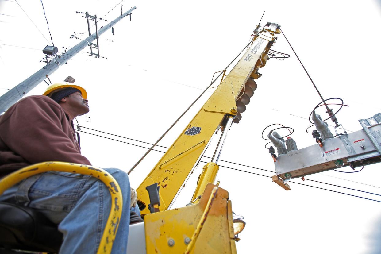 utility worker in jeans sweatshirt and hardhat sits and controls a long arm that's raising a large rectangular metal device toward power lines above