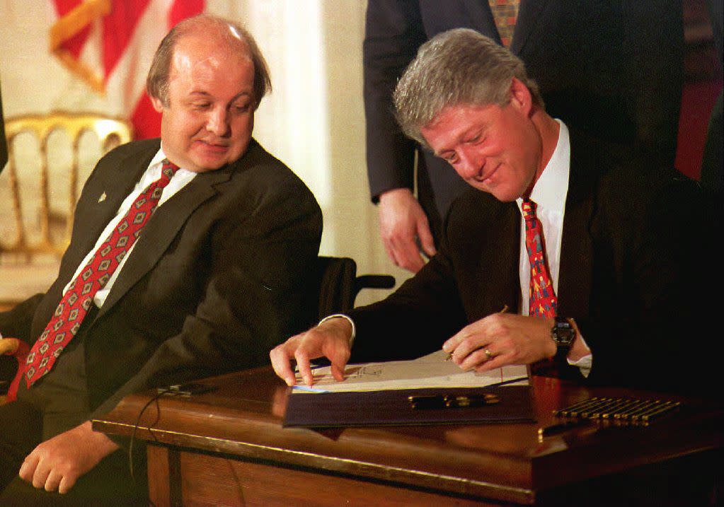 James Brady, seated in a wheelchair, looks over his shoulder as President Bill Clinton signs the Brady bill with a broad smile on his face. 
