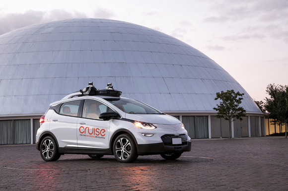 A white Chevrolet Bolt EV with Cruise Automation logos and visible self-driving hardware is parked next to the Design Dome on GM's historic Technical Center campus in Warren, Michigan.