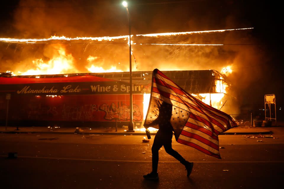 A protester carries the carries a US flag upside, a sign of distress, next to a burning building in Minneapolis.