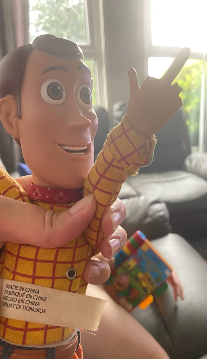 Woody doll from Toy Story being held, showing a tag with manufacturing details. Background includes a window and blurred seating area