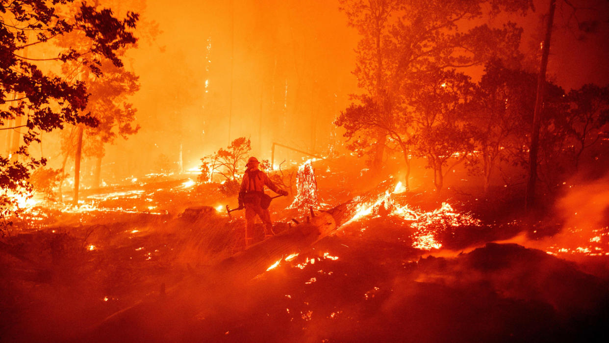 A firefighter carrying an ax in a wooded area against a backdrop of flames as a blaze pushes toward homes.