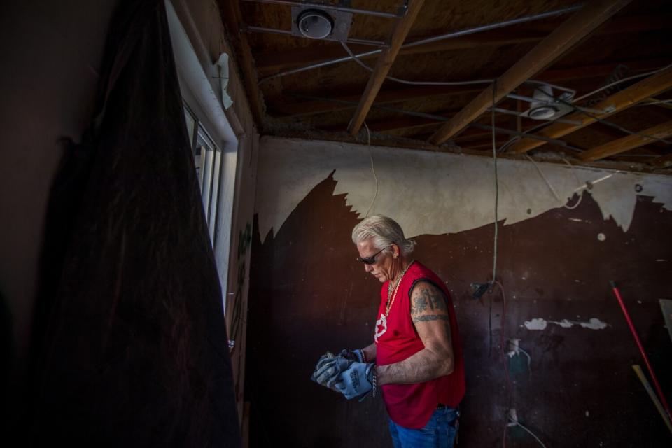 A man in dark glasses, a red top and work gloves looks down while standing in a room with exposed ceiling beams