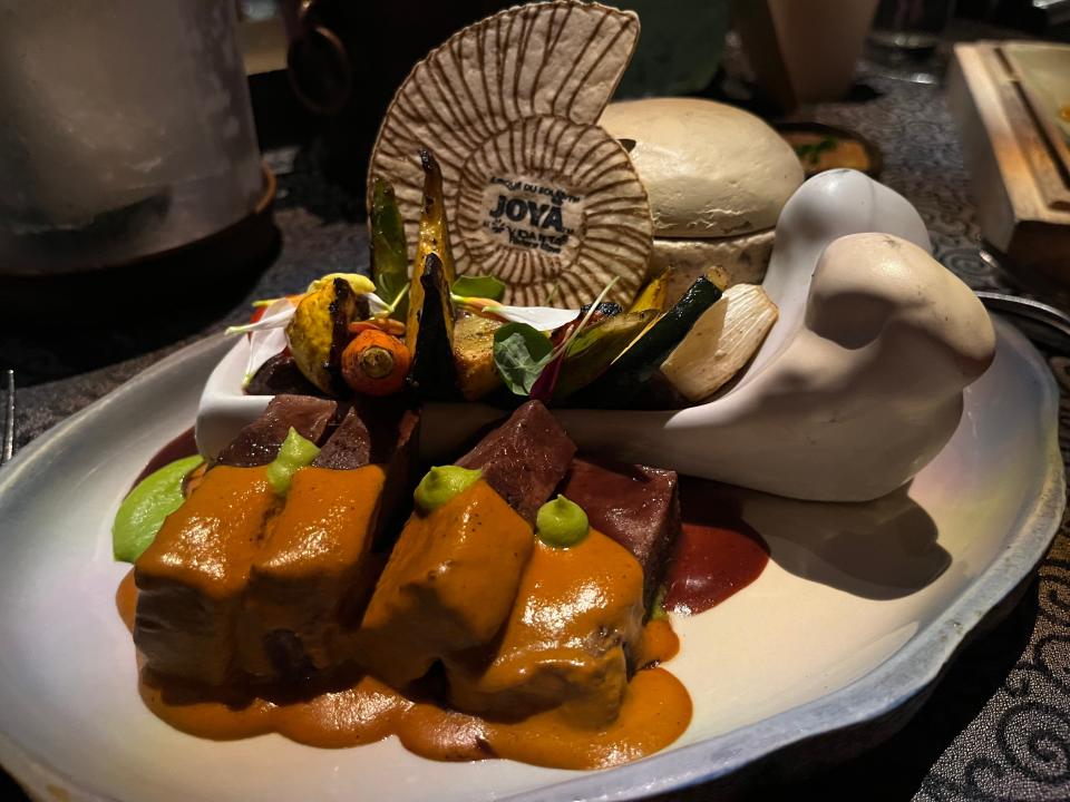 Seashell-like decorative card with Cirque du Soleil Joya logo, short rubs with an orange sauce and green garnish, and vegetables served in a bowl resembling a bone
