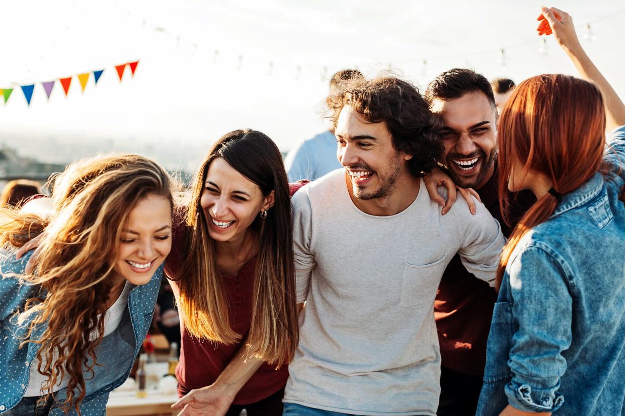 Five young adults having fun and hugging at a rooftop party during the early afternoon, with decorations, other people, and hills blurred in the background