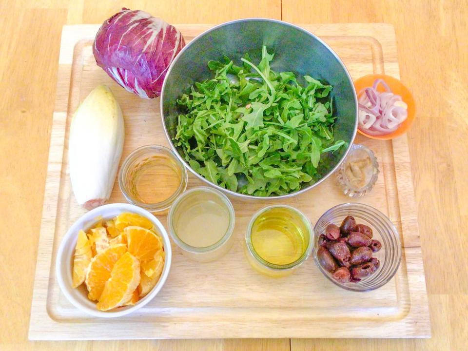A wooden cutting board filled with several bowls of salad ingredients. The bowls contain arugula, shallots, olives, olive oil, cabbage, and orange wedges.