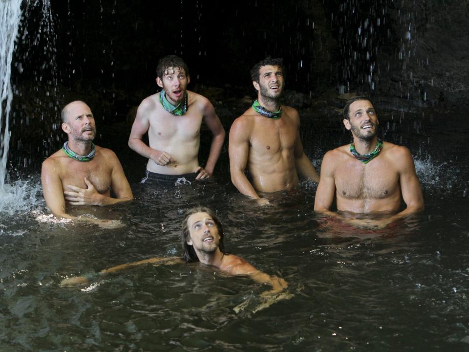 Survivor players standing and sitting in the water by a cave with a waterfall