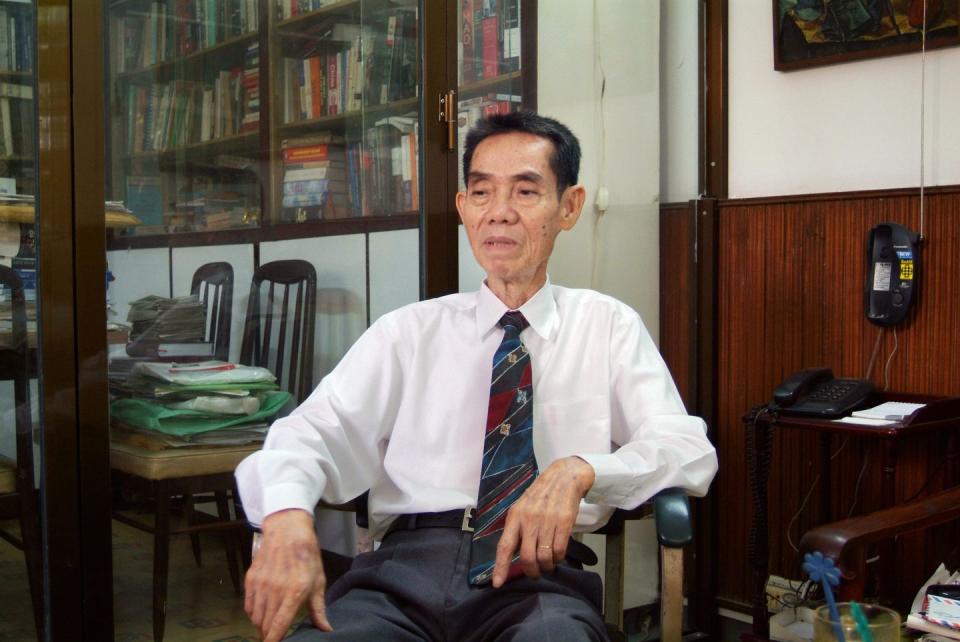 pham xuan an sits in a chair and looks to the left, he wears a white collared shirt, gray slacks, and a blue and red patterned tie