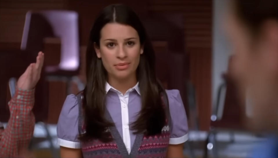 Lea Michele, wearing a white-collared shirt and patterned vest, stands indoors while a person's hand is raised next to her