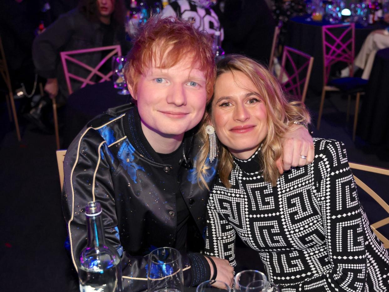 Ed Sheeran (a red-haired man) and Cherry Seaborn (a blonde woman) smiling as they sit at a table.