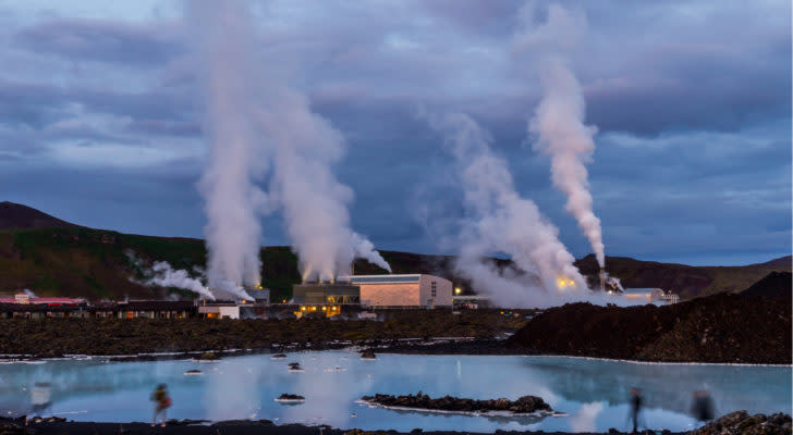 A geothermal power plant operates, with a large lake visible in the foreground and mountainous terrain visible in the background.