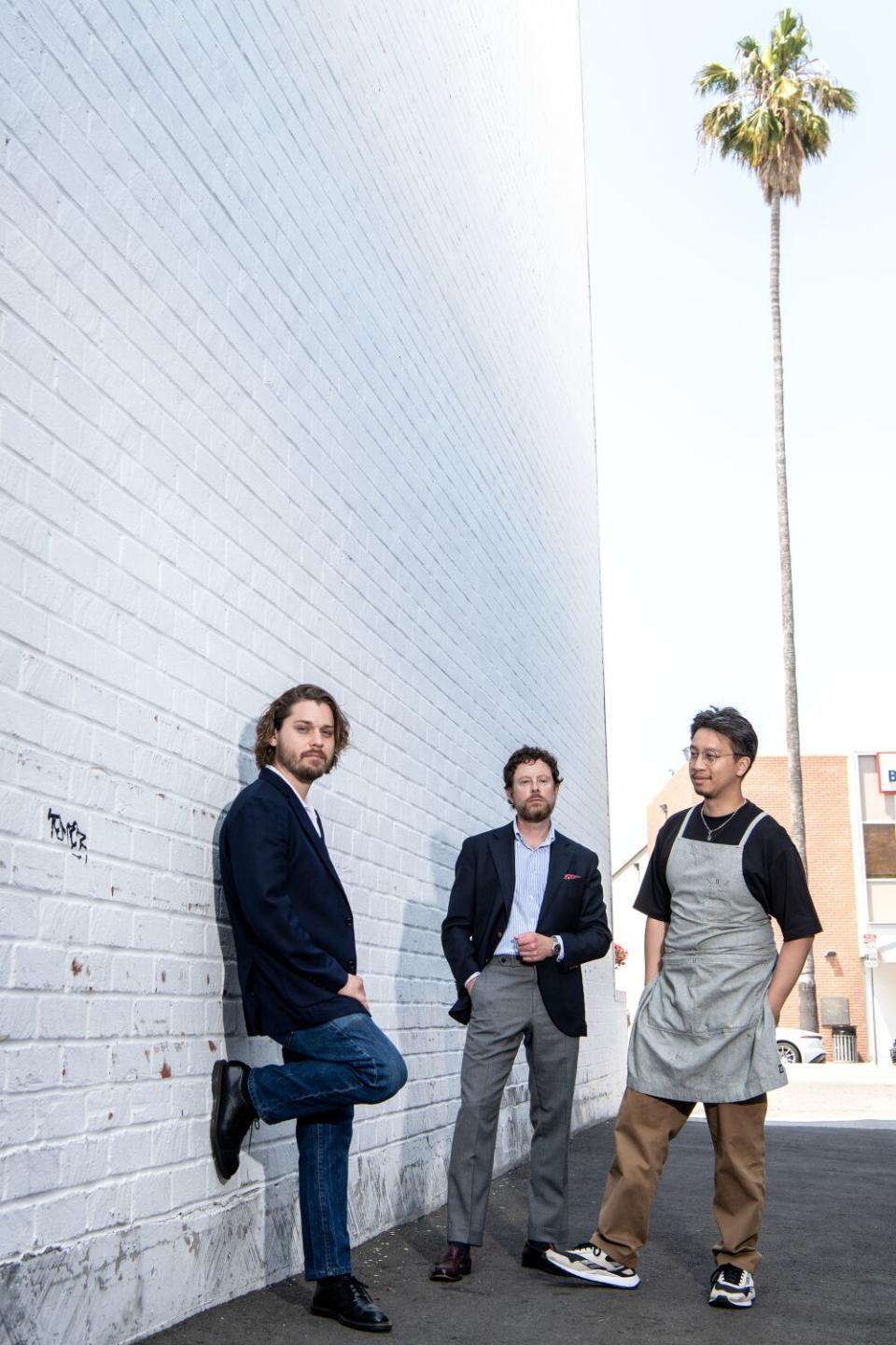 Three men stand in an alley next to a whitewashed brick wall, a palm tree in the distance.