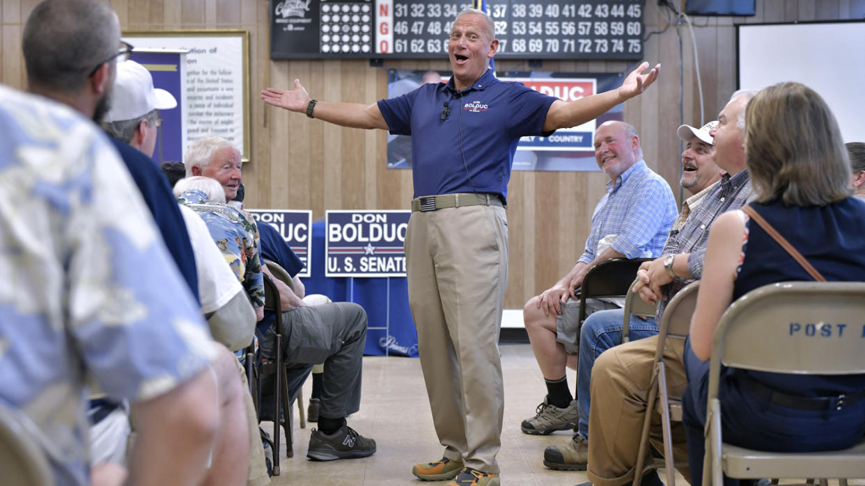 Don Bolduc stands with arms outstretched as people in folding chairs look on. A Bingo board and election signs are seen in the background.