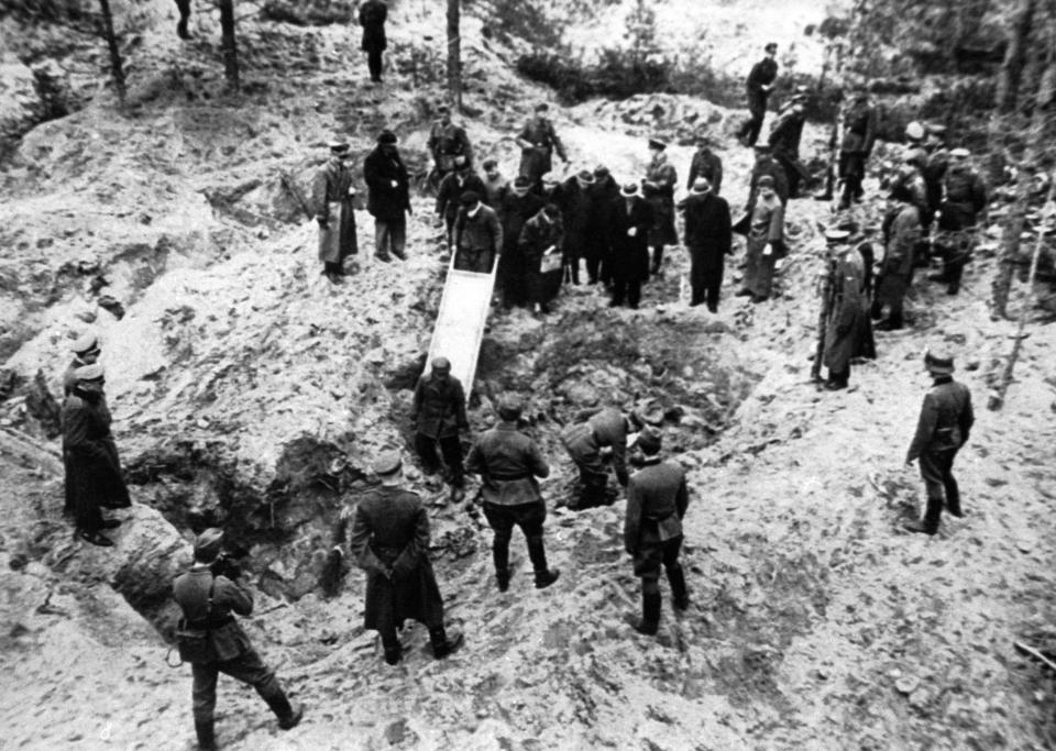 Black and white image, from above, of a mass grave in a clearing with dozens of people wearing long coats and hats or military uniforms standing by as two people hold a stretcher partway into a large hole in the ground.
