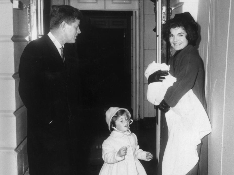 John F. Kennedy smiles as he stands with his wife Jacqueline Kennedy, who holds John Kennedy Jr. in her arms, and their daughter, Caroline, in front of a doorway at the White House in 1961.