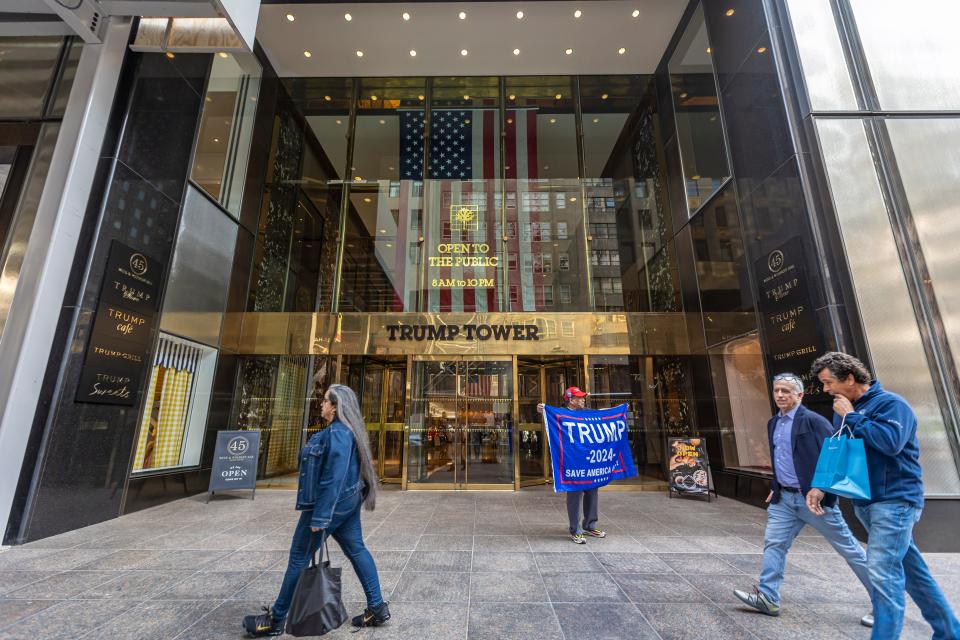 A man holds a "Trump 2024" flag in front of the gold-plated glass facade of the Trump Tower lobby