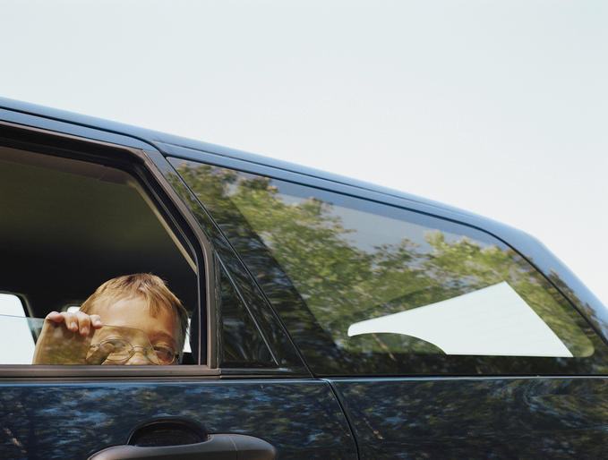 A child looks out of a car window, with their face partially visible and holding a magnifying glass