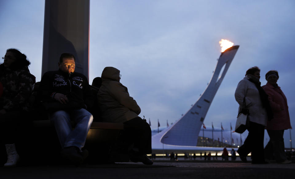 A light from the ground illuminates the face of a visitor to the Olympic Park at dusk as the flame burns in the background at the 2014 Winter Olympics, Thursday, Feb. 20, 2014, in Sochi, Russia. (AP Photo/David Goldman)