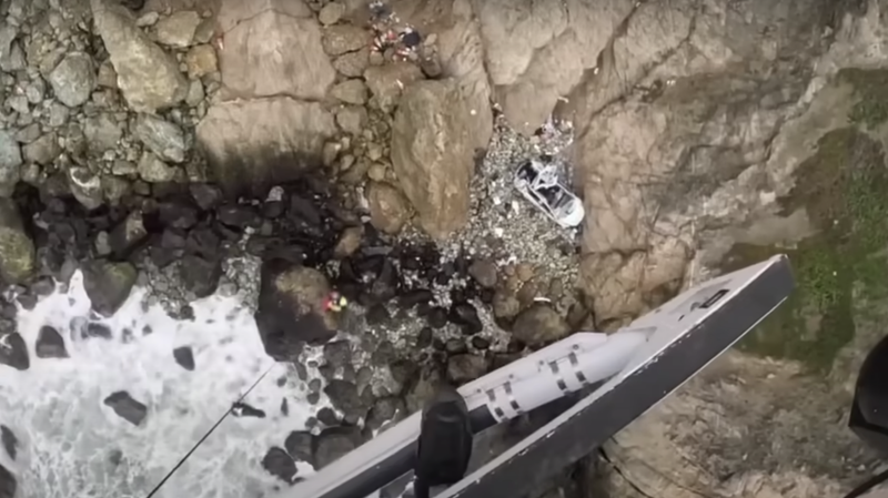 Rescue worker being lowered by rope from a helicopter to the rocky cliffs below where a crashed Tesla Model Y lies on boulders and rocks.