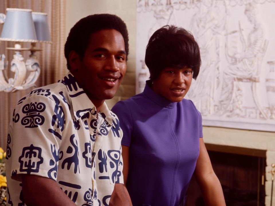 o.j. simpson and marguerite simpson posing together at home and smiling. OJ is in a patterned shirt while marguerite is in a purple short sleeve turtleneck, her hair worn short