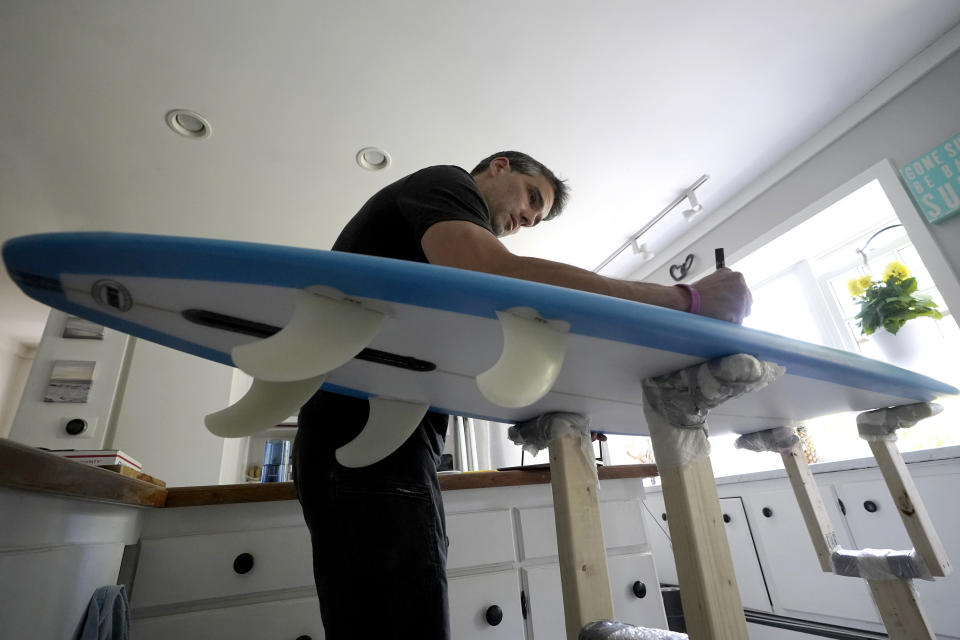 Dan Fischer, of Newport, R.I., uses a paint marker while placing names of lost loved ones on one of his surfboards at his home, in Newport, Wednesday, May 25, 2022. Fischer, 42, created the One Last Wave Project in January 2022 to use the healing power of the ocean to help families coping with a loss, as it helped him following the death of his father. Fischer places names onto his surfboards, then takes the surfboards out into the ocean as a way to memorialize the loved ones in a place that was meaningful to them. (AP Photo/Steven Senne)