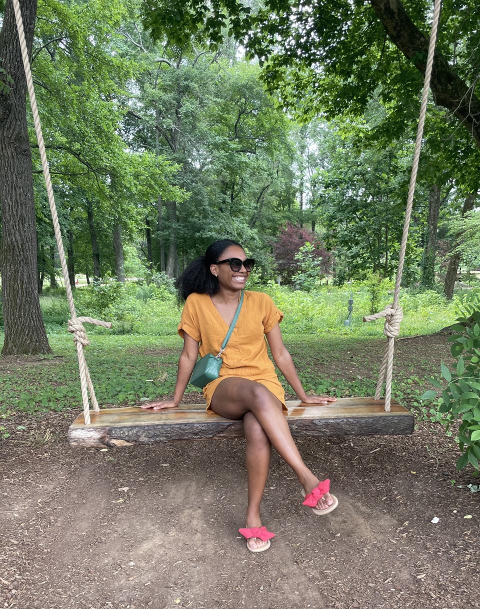 A woman in a casual dress sits on a wooden swing in a forested park, smiling and wearing sunglasses and sandals, with a small crossbody bag