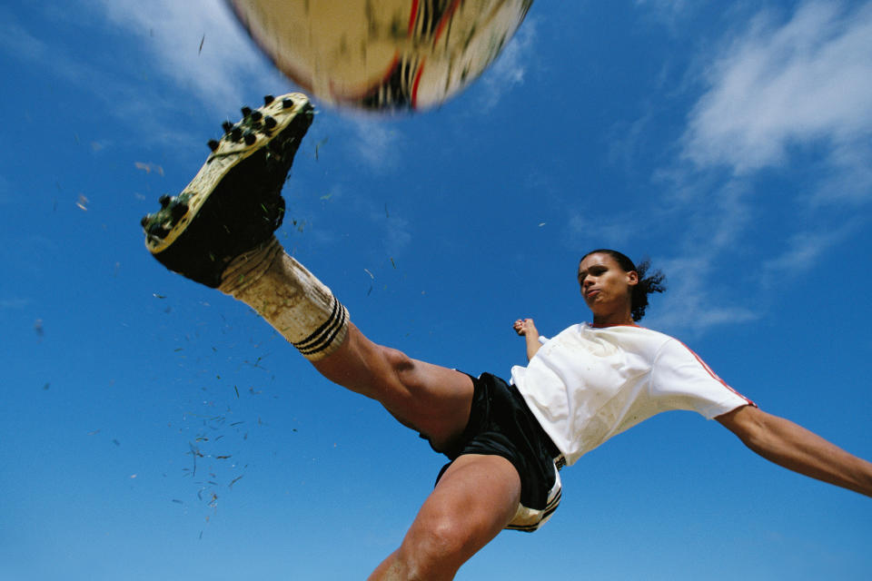 Person kicking a soccer ball mid-air, shot from a low-angle perspective against a clear sky