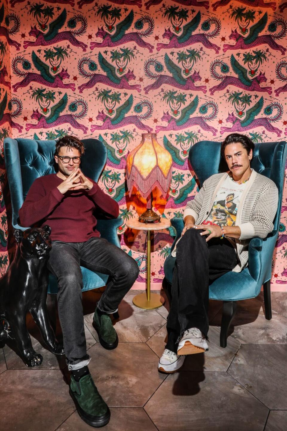 Tom Schwartz and Tom Sandoval sit in velvet wingback chairs in front of pink patterned wallpaper.