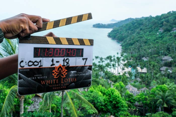 A pair of hands holding a clapperboard for "The White Lotus" in front of a lush coastal view