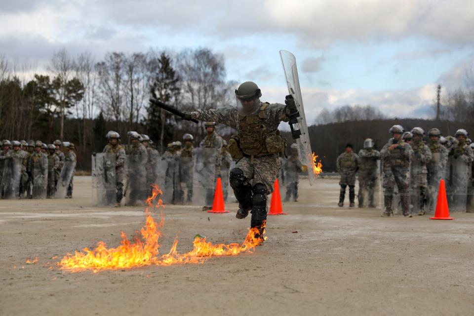 A photo of a man in army gear with his arms out to his sides, with a baton in his right hand and a riot shield in his left. He stands on one foot while the other is prepared to stomp down.