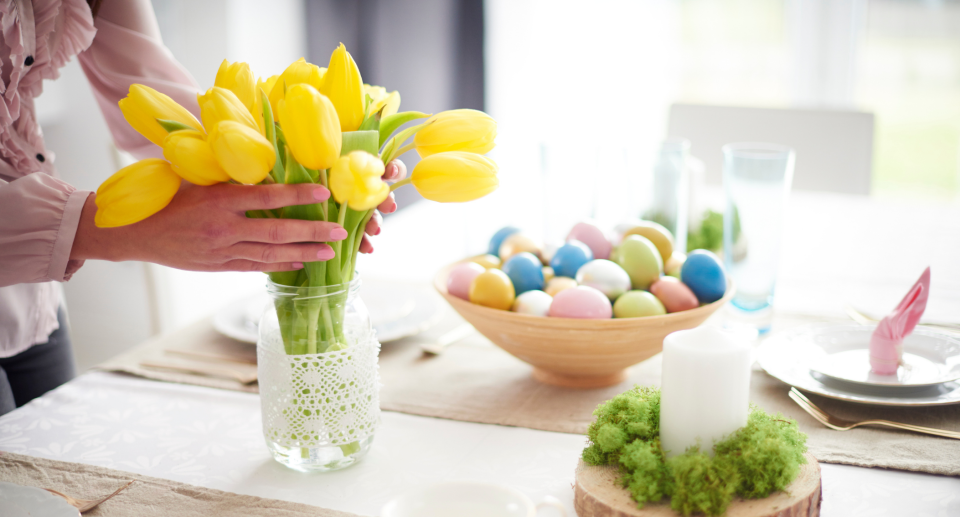 woman putting yellow tulips in vase on table with easter eggs and candle