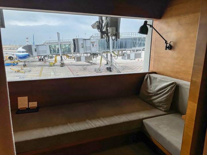 A cozy sleep suite is shown at Cathay Pacific's The Pier, a first- class lounge at Hong Kong International Airport. The simple nook has gray cushions and a pillow and a window that overlooks the airport tarmac.