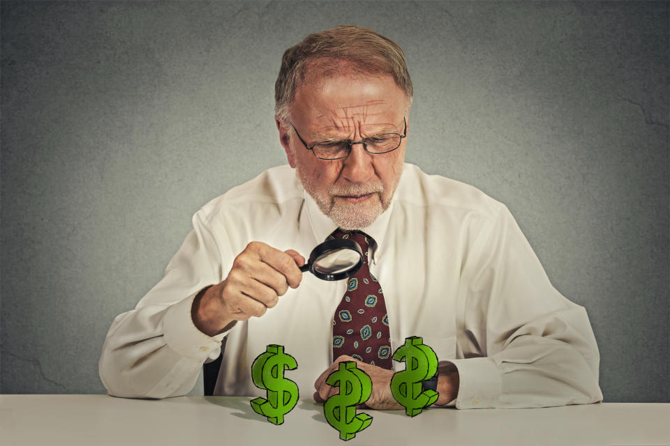 A senior man holding a magnifying glass while examining dollar signs on the counter in front of him.