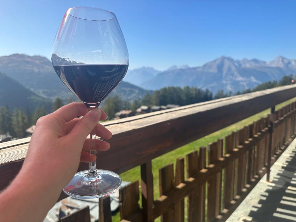 hand holding glass of red wine in front of a wooden railing on a deck overlooking the swiss alps mountain peaks