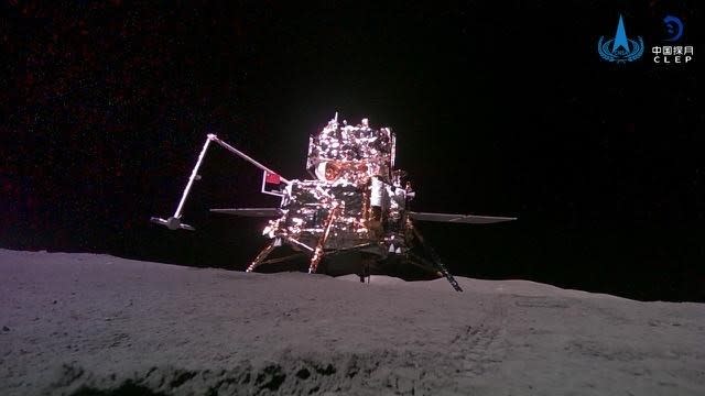  A robotic lander stands on a barren grey surface against a black sky. 