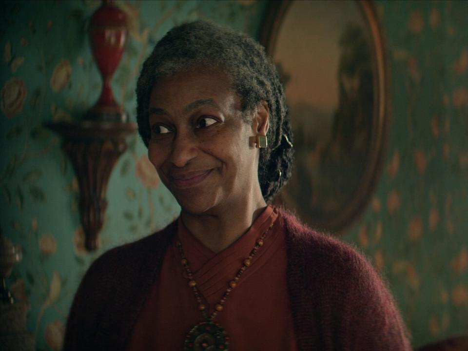 sandra james-young as unity kincaid in the sandman. she's an older woman with grey hair drawn back into a bun, wearing warm colors and smiling while looking towards the side.