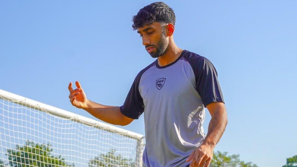 Imran Kayani looking down at a football which is not visible in the photo because it is cropped to his waist. He is British Pakistani and is wearing a grey and navy T-shirt that says PFF (Pakistani football federation) on the front. He has short black hair and a trimmed black beard and moustache. Behind him you can make out the top of a football goal and the net and there is a clear blue sky.