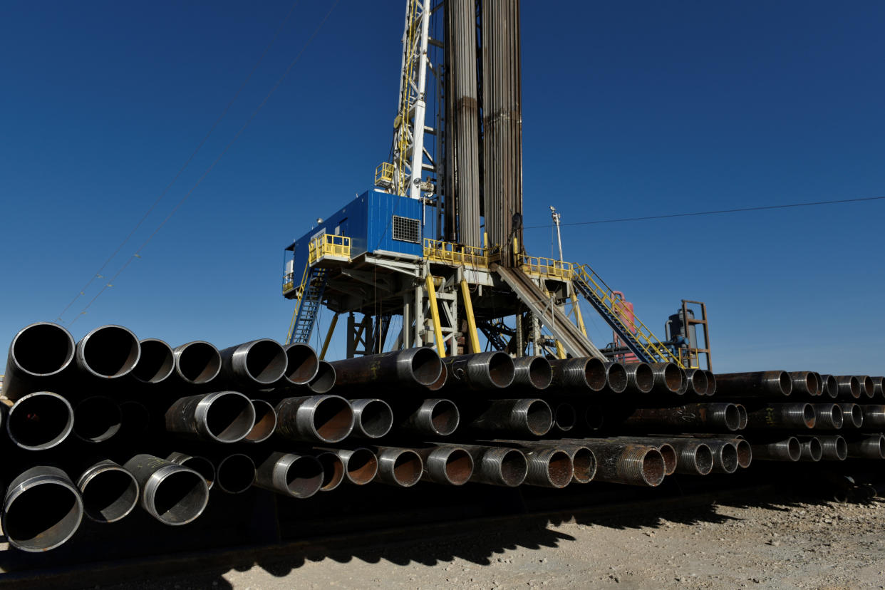 More than 50 large-diameter pipes with threaded ends stacked against a blue sky and drilling equipment.