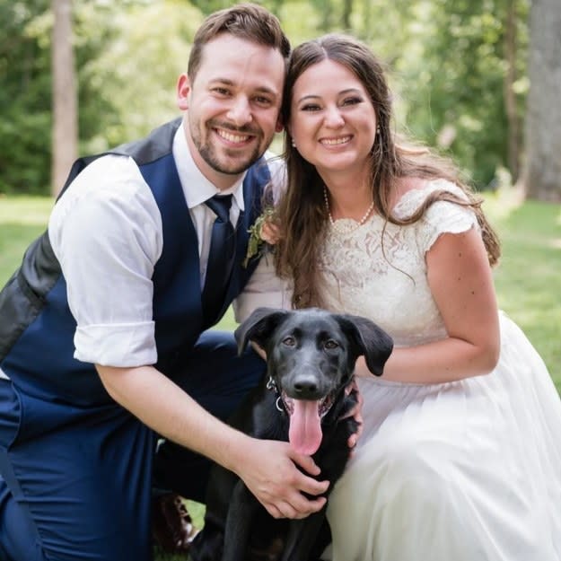 Bucky standing and smiling in between his owners during the wedding