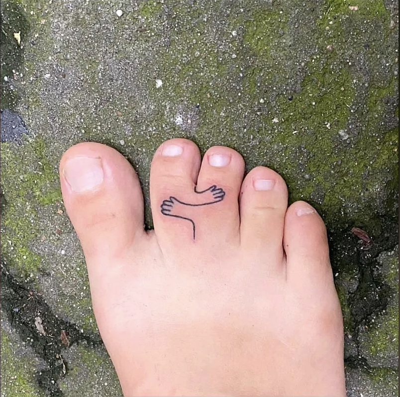 Toe with a whimsical tattoo of a small arm sticking out, appearing to grasp the second toe. Green mossy ground in background