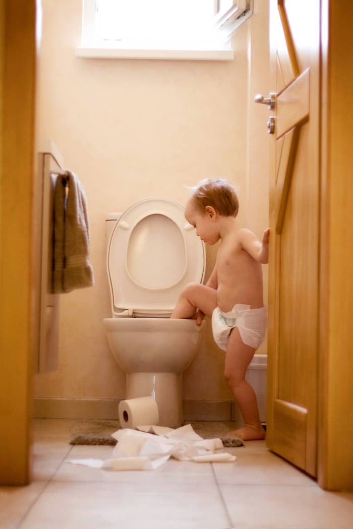 A baby in a diaper is standing next to a toilet with one foot inside it. Toilet paper is strewn on the bathroom floor