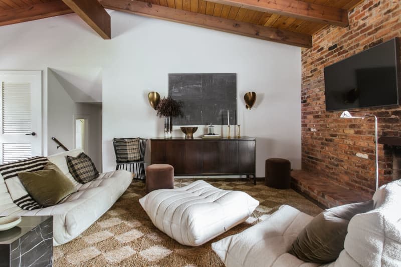 Red wood ceiling with large beams, brick accent wall with tv, brown and cream checkered rug, soft white modern shaped couches